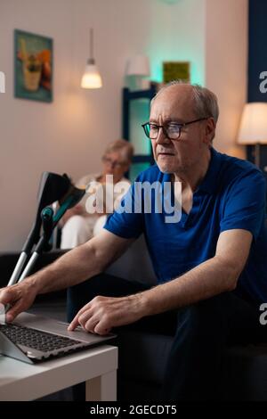 Personnes âgées en retraite dans la salle de séjour à la maison. Femme caucasienne en fauteuil roulant de lecture et homme vieux regardant le périphérique virtuel ordinateur portable Internet avec des béquilles Banque D'Images