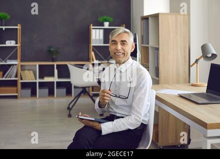 Homme d'affaires senior heureux assis sur une chaise dans son bureau, tenant des lunettes et souriant Banque D'Images
