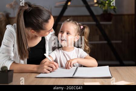 Jeune mère et petite fille caucasiennes assis à la table le soir, étudier en ligne ensemble, mère et petite fille écrire à la main, faire des devoirs, Banque D'Images