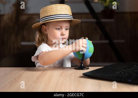 Une étudiante de race blanche vêtue d'un chapeau tenant un globe dans ses mains est assise à la maison à une table avec un ordinateur portable. Banque D'Images