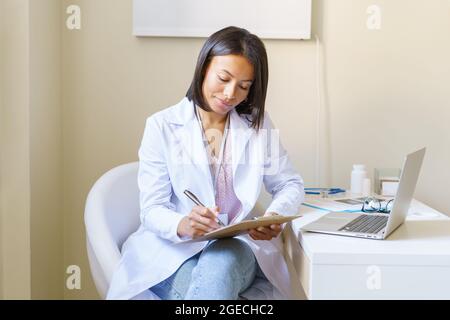 Jeune femme africaine médecin en manteau blanc assis sur le lieu de travail en clinique et remplissant la forme du patient Banque D'Images