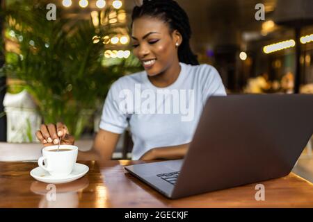 Portrait d'une jeune femme africaine charmante blogueuse, dactylographiant un nouveau poste stimulant pendant le déjeuner dans un café, avec ordinateur portable et smartphone avec copie s. Banque D'Images