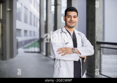 Portrait horizontal à la taille d'un beau médecin malais arabe, portant un manteau blanc, debout avec les bras croisés à l'extérieur sur l'arrière-plan de la clinique Banque D'Images