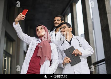 Groupe de trois jeunes médecins stagiaires ou étudiants en médecine prenant des selfies tout en se tenant à l'extérieur de l'hôpital. L'équipe médicale de Mutliethnic fait du selfi Banque D'Images