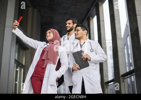 Groupe de trois jeunes médecins stagiaires ou étudiants en médecine prenant des selfies tout en se tenant à l'extérieur de l'hôpital. L'équipe médicale arabe fait le pH du selfie Banque D'Images