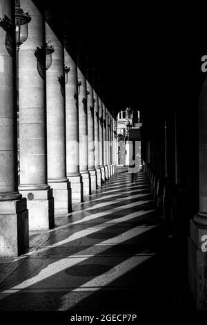 Passerelle parisienne en noir et blanc. Une personne non identifiable marche dans les ombres projetées par les anciennes colonnes architecturales. Paris, France. Banque D'Images