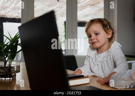 Petite fille mignonne à écouter l'école en ligne tout en regardant sur un ordinateur portable dans la cuisine. Banque D'Images