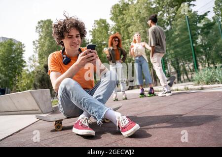 jeune homme souriant assis sur un skateboard et messagerie sur un smartphone à proximité de patineurs interraciaux flous Banque D'Images