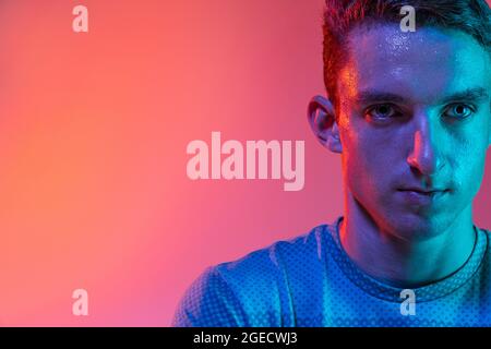 Gros plan jeune homme sportif caucasien debout sur fond de studio rose avec filtre néon bleu, lumière. Sens du but Banque D'Images