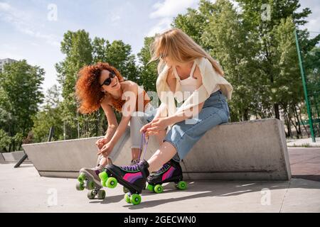 femme gaie en lunettes de soleil nouant des lacets sur le patin à roulettes sur la frontière près d'un ami Banque D'Images