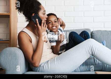 Femme noire parlant au téléphone portable tout en embrassant son fils à la maison Banque D'Images