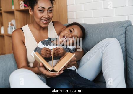 Mère et fils souriants noirs lisant le livre tout en étant assis sur un canapé à la maison Banque D'Images