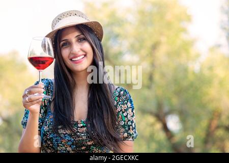 Jeune femme heureuse avec chapeau de paille et verre de vin rouge, en arrière-plan Banque D'Images