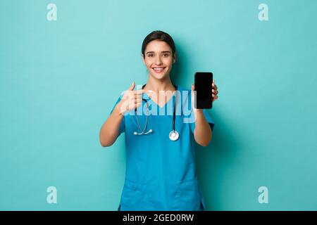 Covid-19, concept de pandémie et de médecine. Souriant médecin souriant dans des exfoliations, pointant le doigt vers le téléphone mobile, montrant l'application, debout sur bleu Banque D'Images