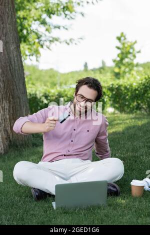 Homme d'affaires joyeux tenant une carte de crédit pendant un appel vidéo sur un ordinateur portable sur l'herbe Banque D'Images