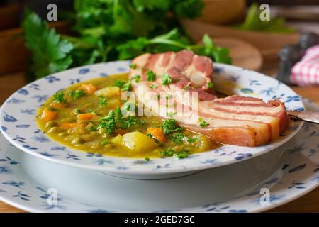 Soupe maison aux pois verts avec légumes-racines, pommes de terre et poitrine de porc servie sur une table rustique en bois Banque D'Images