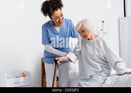 Une infirmière afro-américaine souriante aidant le patient à marcher la canne près du lit d'hôpital Banque D'Images