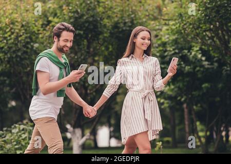 Photo portriat jeune couple souriant marchant dans le parc vert tenant les mains à l'aide de smartphones Banque D'Images