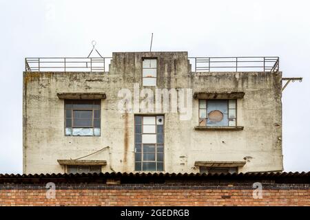 Un bâtiment délabré avec des fenêtres boardées et cassées se dresse isolé derrière un mur de briques. Banque D'Images