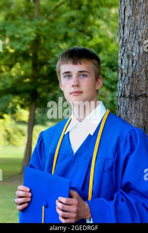 Le jeune homme porte sa robe de graduation bleue et tient son chapeau. L'or décorera ses épaules. Il est debout à l'extérieur et solennel. Banque D'Images