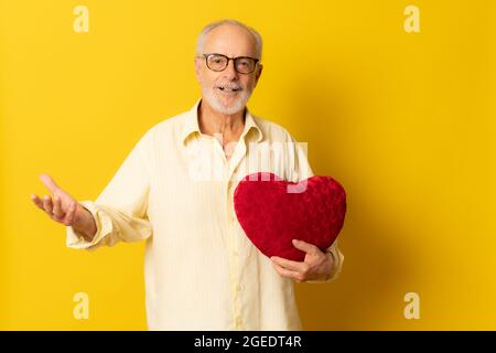 Homme âgé tenant un oreiller en forme de cœur sur fond jaune. Banque D'Images