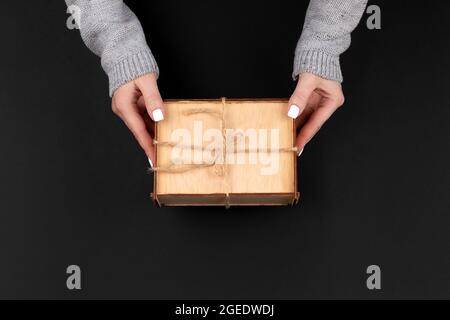Femme mains en tricot gris chandail avec ongles blancs tenant en bois rustique boîte cadeau décorée avec corde de chanvre sur fond noir. Vue de dessus, plan de travail c Banque D'Images