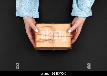 Femme mains en tricot bleu chandail avec ongles blancs tenant en bois rustique boîte cadeau décorée avec corde de chanvre sur fond noir. Vue de dessus, plan de travail c Banque D'Images