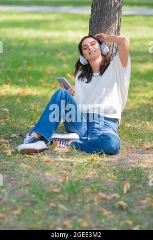 Photo d'une femme de 40 ans dans un casque avec un smartphone. Elle ferme les yeux et écoute sa musique préférée tout en étant assise sur l'herbe dans le Banque D'Images