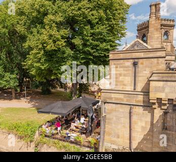 Un café avec des parasols se trouve dans un parc public et à côté d'une rive. Une tour se dresse au-dessus et fait partie d'un pont historique et orné. Banque D'Images