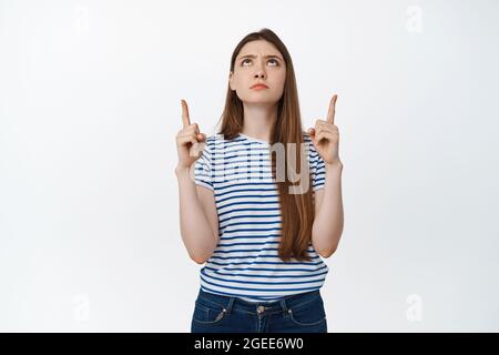 Jeune femme pointant les doigts vers le haut et frowning douteux, regardez le logo déçu, montrant quelque chose de mauvais, se tenant sur fond blanc Banque D'Images