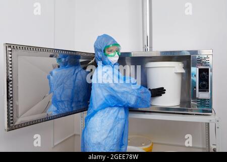 Technologue portant un vêtement de protection EPI, un masque, des gants et travaillant avec un grand seau en plastique dans un laboratoire de recherche Banque D'Images