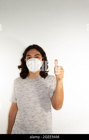 Homme hispanique aux cheveux longs portant un masque de kn95 sur fond blanc avec un pouce vers le haut Banque D'Images