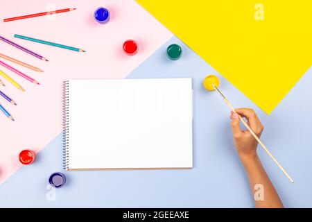 Main d'enfant avec peinture au pinceau dans carnet de croquis d'art vierge. Vue de dessus de l'art créatif fournitures aquarelles crayons sur la table. Retour à Banque D'Images