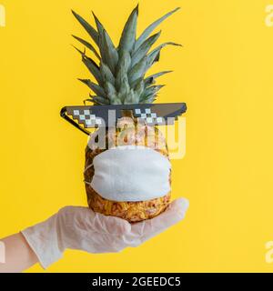 Visage d'ananas drôle dans un masque médical de protection. La main du médecin dans un gant en caoutchouc contient l'ananas aux fruits tropicaux d'été dans des lunettes de soleil. Concept de sécurité de voyage Banque D'Images