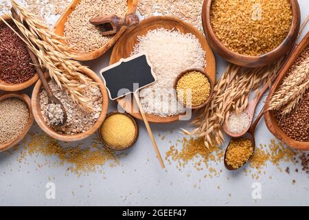 Riz blanc, brun et rouge, sarrasin, millet, gruaux de maïs, quinoa et bulgur dans des bols en bois sur la table de cuisine gris clair. Céréales sans gluten. Haut Banque D'Images