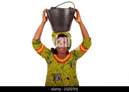 belle femme adulte africaine vêtue de tissu vert, équilibrant un seau noir sur sa tête avec ses mains, souriant Banque D'Images