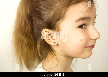 Perçage d'oreilles chez un enfant - une fille montre un anneau auriculaire dans son oreille fait d'un alliage médical. Fond blanc, portrait d'une fille avec une taupe sur sa joue Banque D'Images