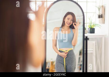 La jeune femme mesure sa taille avant le miroir avec un geste OK. Fitness. Banque D'Images