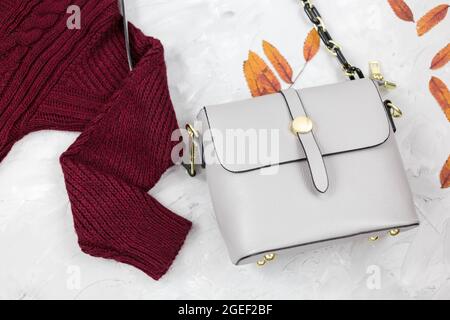 Sac femme en cuir gris, chandail rouge foncé, feuilles d'automne sur fond gris. Accessoires tendance pour l'automne. Tenue confortable Banque D'Images