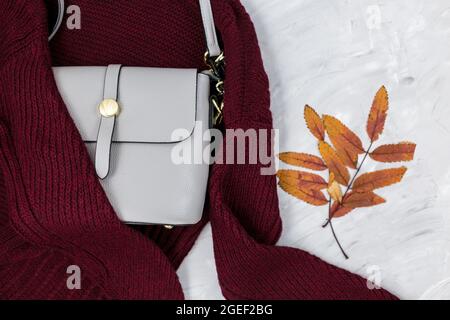 Sac femme en cuir gris, chandail rouge foncé, feuilles d'automne sur fond gris. Accessoires tendance pour l'automne. Tenue confortable Banque D'Images