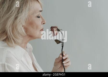 Triste femme âgée senior en chemise blanche tient avec une fleur rose grise vieille mort sèche. Vieux concept de vieillissement. Banque D'Images