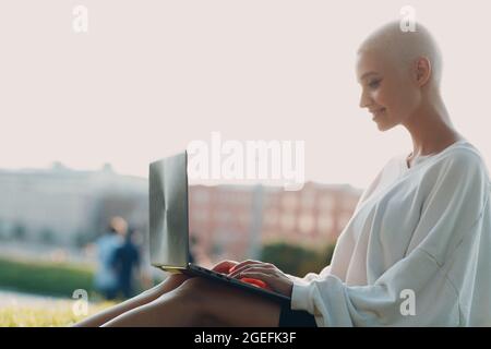 Portrait d'une jeune femme européenne à poil court souriante utilisant un ordinateur portable dans une prairie d'herbe verte dans le parc. Belle fillette blonde heureuse en plein air. Vêtements d'été pour femmes. Banque D'Images