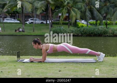 Posez la femme dans des vêtements de sport roses et des sneakers blanches faisant de l'exercice de planche dans le parc avec l'ordinateur portable. Sport en ligne Banque D'Images