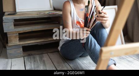 École d'artistes. Passe-temps créatif. Femme assise sur le sol montrant la peinture pinceau éclaboussures colorées disponibles sans visage. Banque D'Images