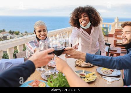 Les jeunes gens multiraciaux applaudissent avec le vin et mangent avec des masques sous les chins au restaurant patio - Focus sur le visage de la femme africaine Banque D'Images