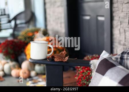 Tasse de café à la vapeur assise sur le bras de la chaise à bascule sur un porche avant qui a été décoré pour l'automne avec des citrouilles blanches, orange et grises Banque D'Images