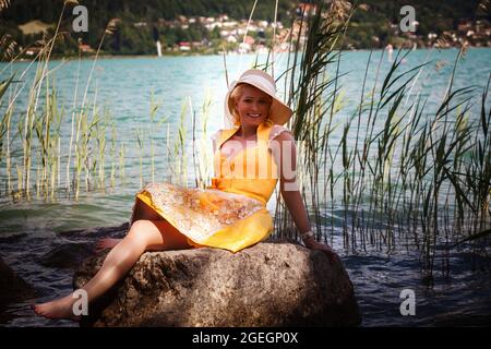 Femme élégante avec dirndl et chapeau de soleil se trouve sourire sur un rocher dans un lac Banque D'Images