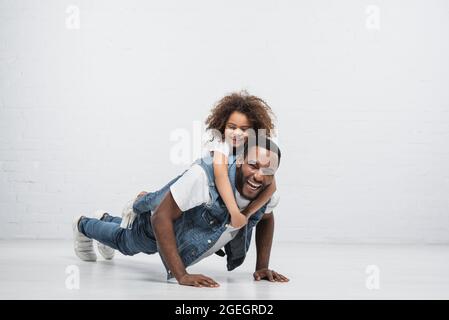 joyeux homme afro-américain qui fait des retouches avec une fille heureuse qui le soutient sur le gris Banque D'Images