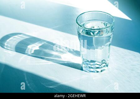 Un verre avec de l'eau claire et claire et des ombres vives se tient sur une table blanche Banque D'Images