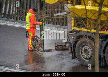 Entretien dans un trou d'homme avec un homme méconnaissable, travaillant derrière le véhicule de nettoyage d'égout sur la rue ouverte. Banque D'Images